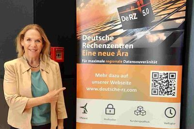 Susanne Back steht neben einem Werberollout der DeRZ mit Informaitonen über das Produkt DeRZ 5.0